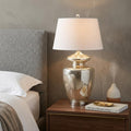 31" Silver Novelty Table Lamp With Drum Shade displayed on a dark wood tabletop in a furnished interior against a tan wall.