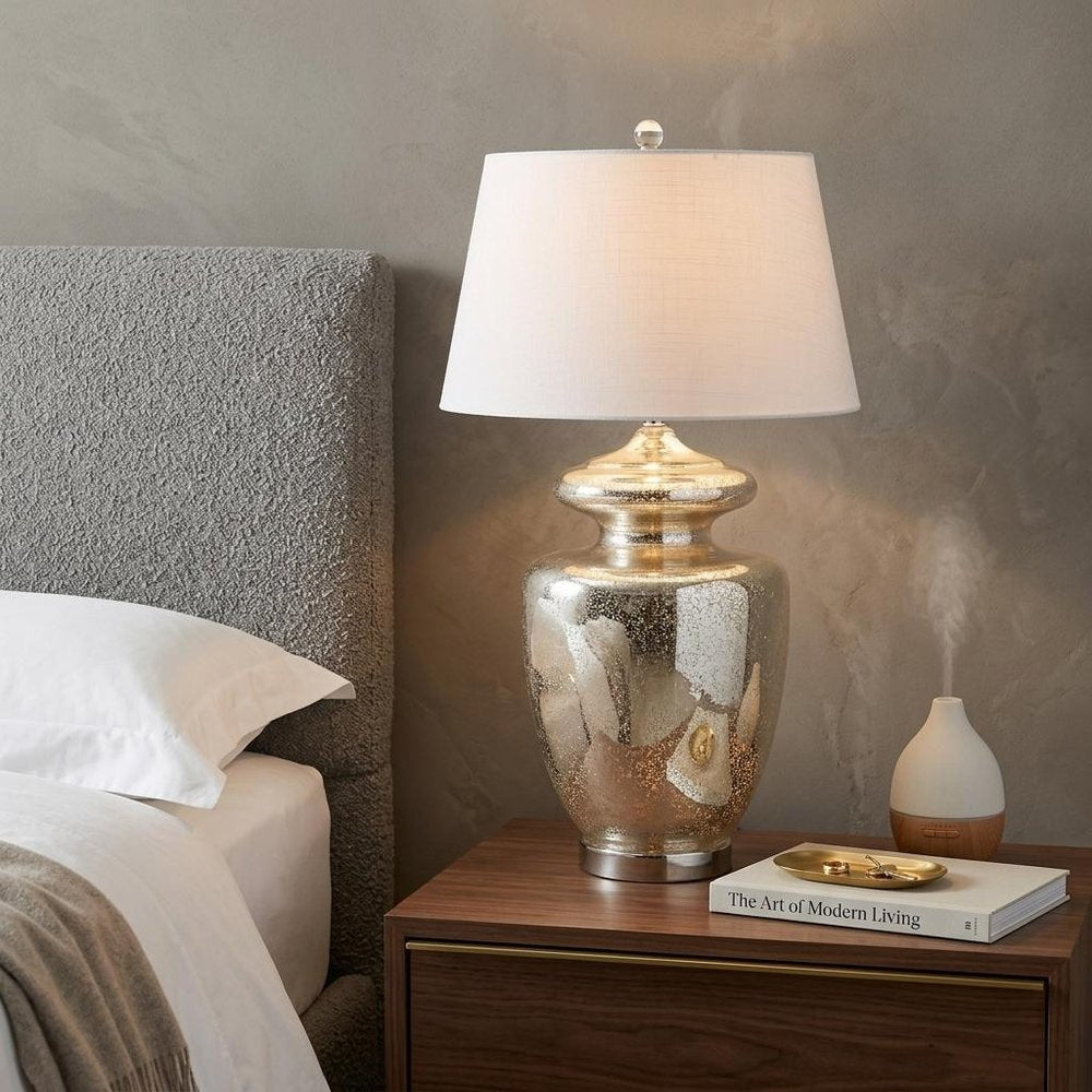31" Silver Novelty Table Lamp With Drum Shade displayed on a dark wood tabletop in a furnished interior against a tan wall.