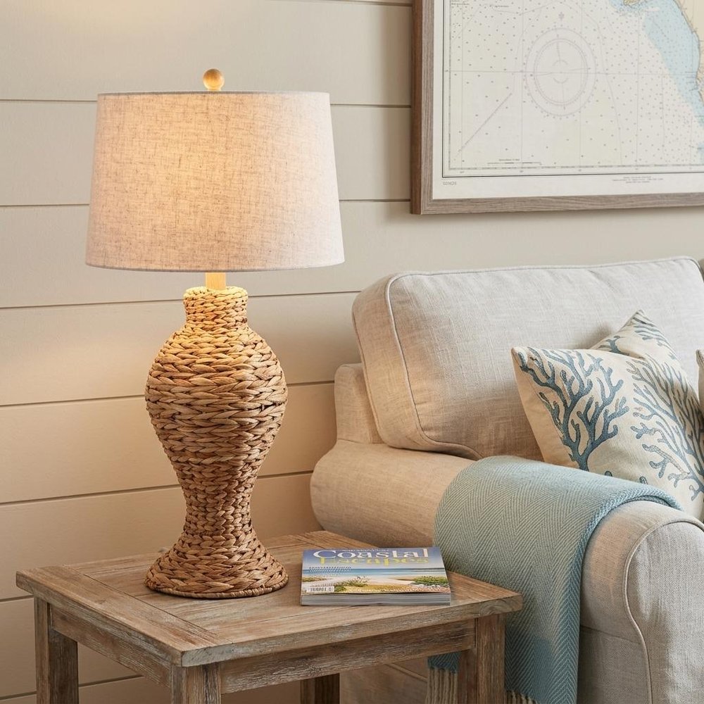 31" Natural Water Hyacinth Urn Table Lamp With Drum Shade displayed on a dark wood tabletop in a furnished interior against a cream wall in tan.