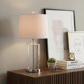 25" Silver Bedside Lamp With Drum Shade styled in a furnished interior with a tan wall on a dark wood floor.