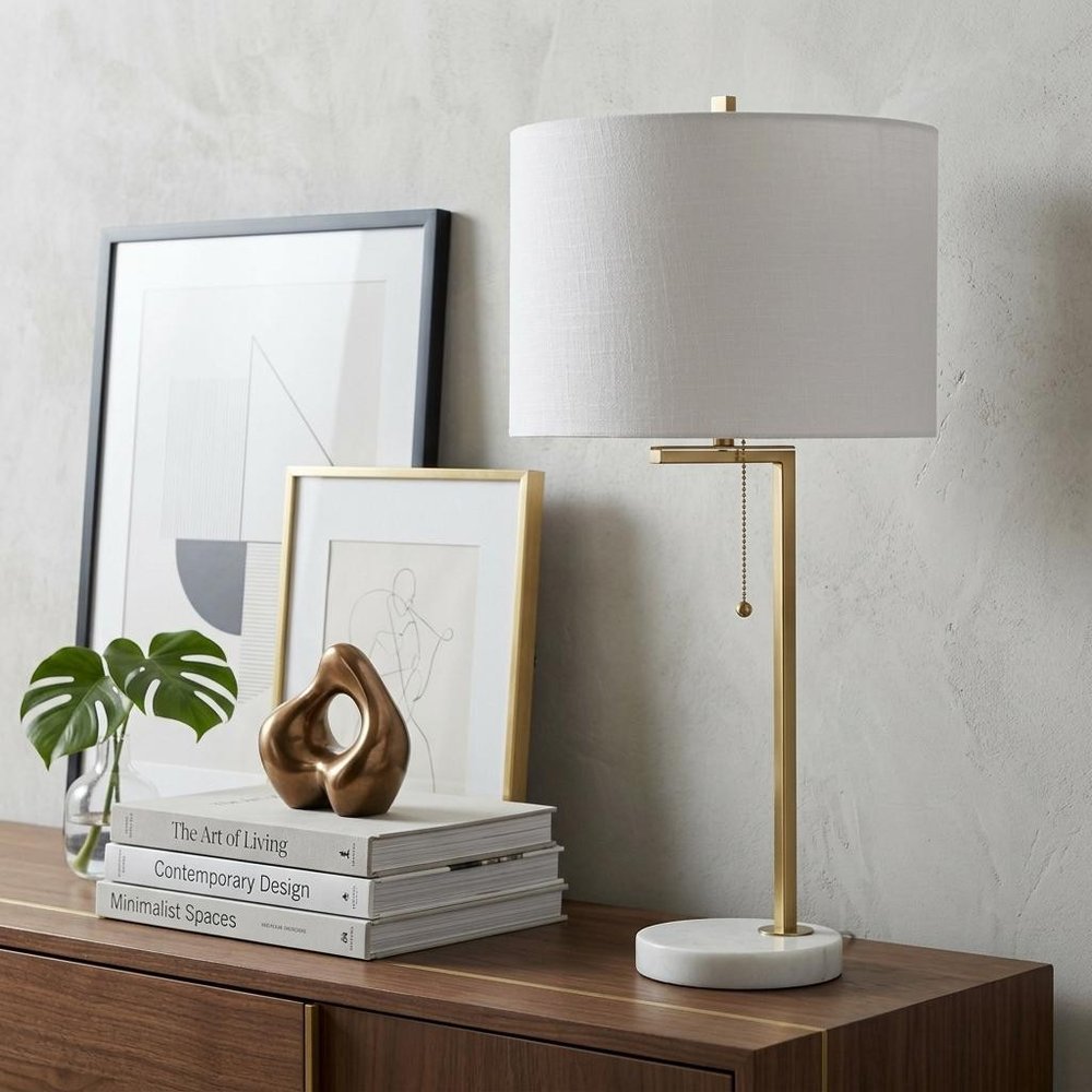 25" Brass And White Round Table Lamp With Drum Shade displayed on a dark wood tabletop in a furnished interior against an off-white wall.