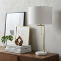 25" Brass And White Round Table Lamp With Drum Shade displayed on a dark wood tabletop in a furnished interior against an off-white wall.
