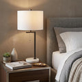 25" White Novelty Table Lamp With Drum Shade displayed on a dark wood tabletop in a furnished interior against a brown wall.
