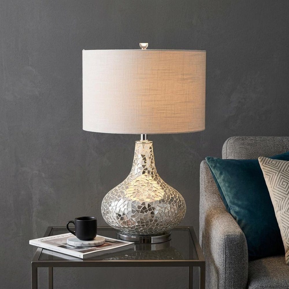 26" Silver Novelty Table Lamp With Drum Shade displayed on a tabletop in a furnished interior against a dark wall.