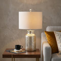 25" Clear Table Lamp With Drum Shade displayed on a dark wood tabletop in a furnished interior against a tan wall.
