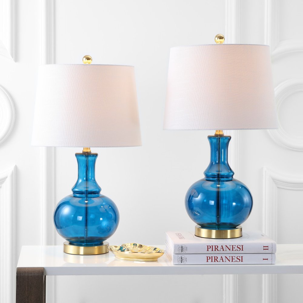 Two blue glass table lamps with white drum shades and gold bases on a white surface with a white wall background.