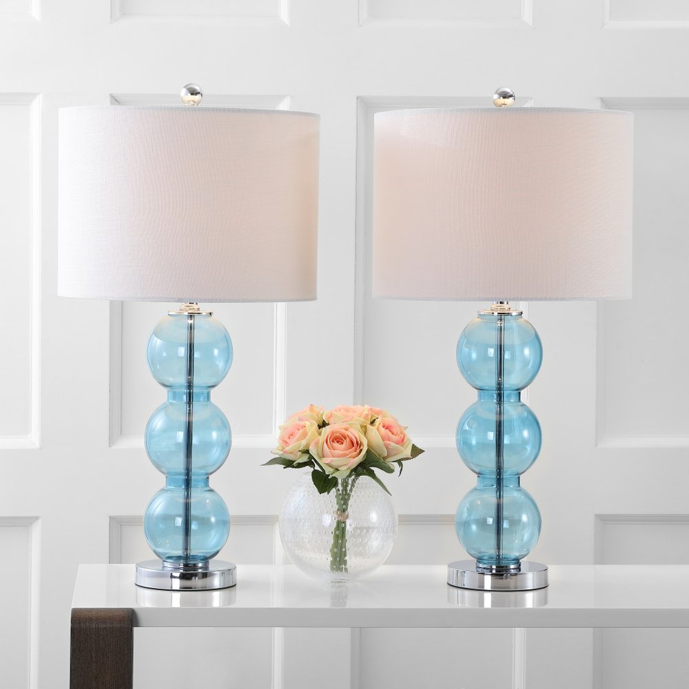 Two blue glass ball table lamps with white drum shades on a white table in front of a white paneled wall.