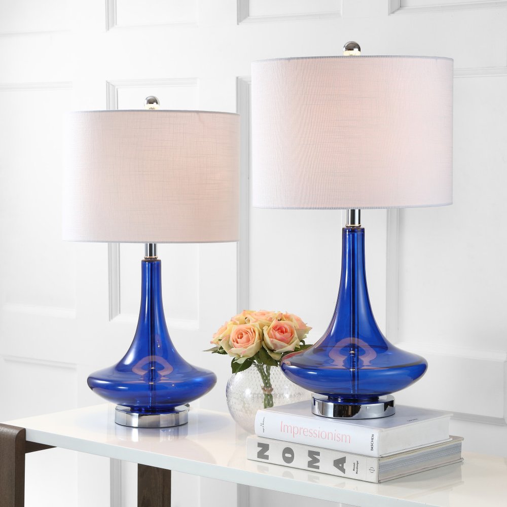 Two blue glass table lamps with white drum shades on a white table with books and flowers in the background.