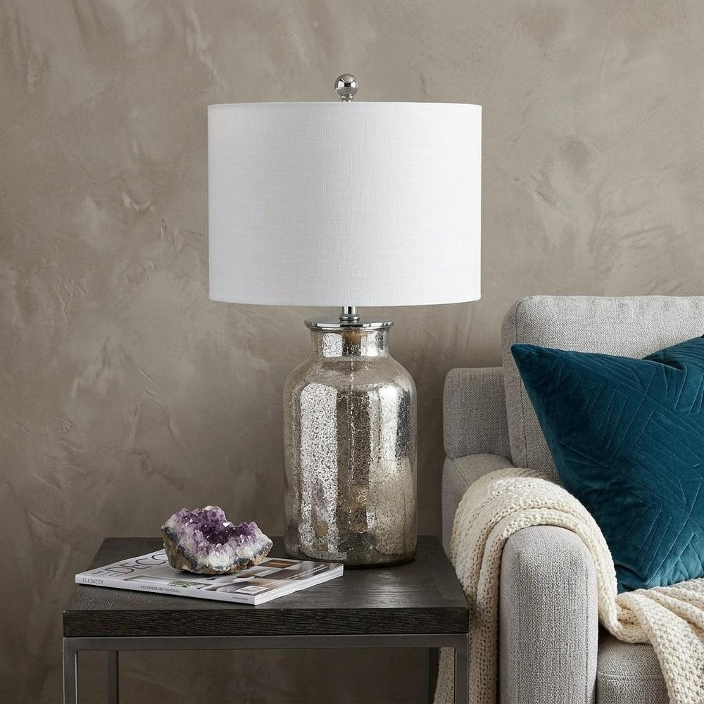 25" Clear Novelty Table Lamp With Drum Shade displayed on a dark wood tabletop in a furnished interior against a tan wall.