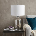 25" Clear Novelty Table Lamp With Drum Shade displayed on a dark wood tabletop in a furnished interior against a tan wall.