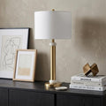 27" Gold Novelty Table Lamp With Drum Shade displayed on a tabletop in a furnished interior against a tan wall.