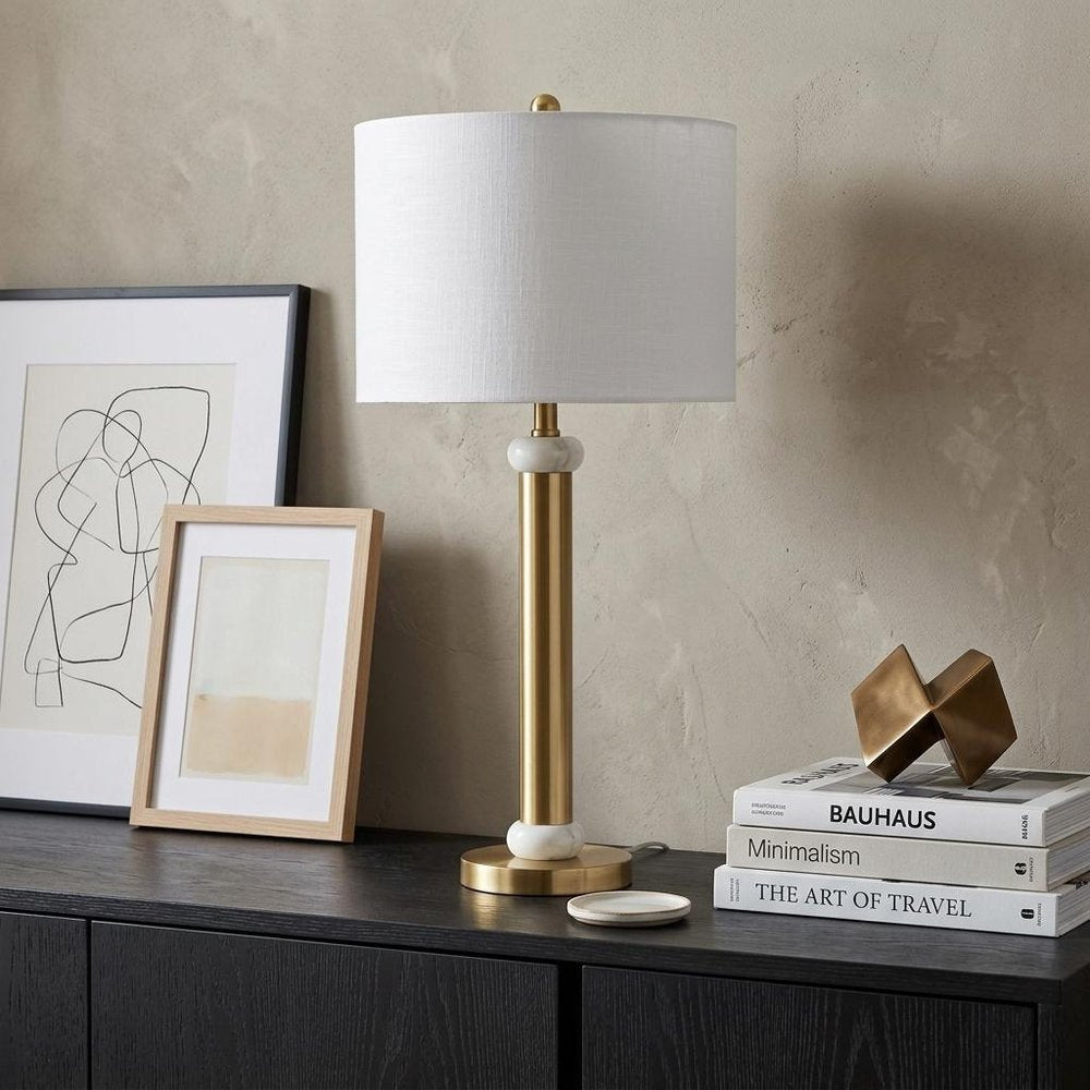 27" Gold Novelty Table Lamp With Drum Shade displayed on a tabletop in a furnished interior against a tan wall.