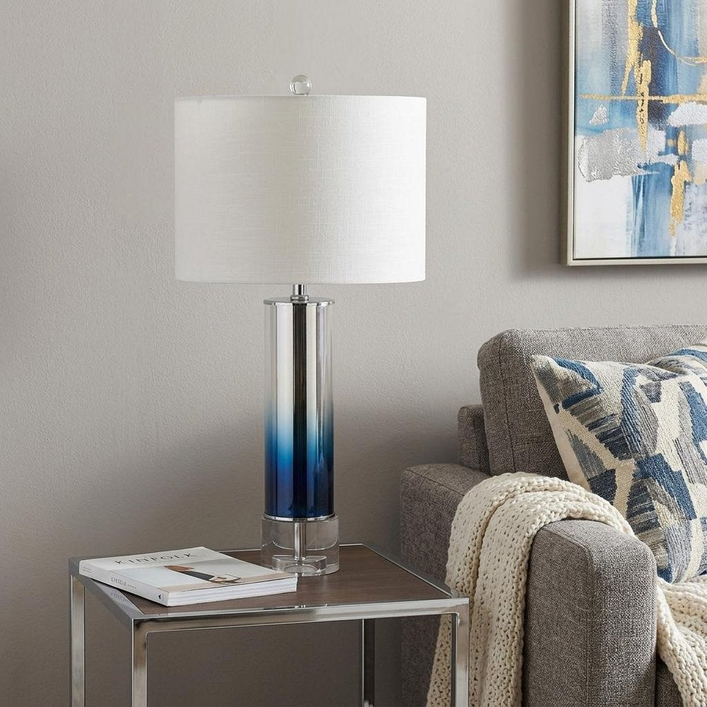 27" Clear Novelty Table Lamp With Drum Shade displayed on a dark wood tabletop in a furnished interior against a light gray wall.