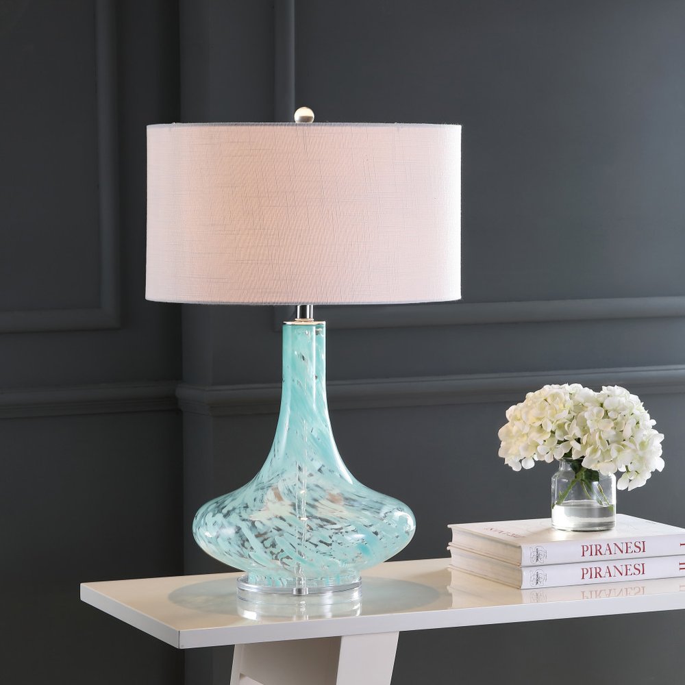 29" Light Blue And Clear Teardrop Glass Table Lamp With Drum Shade displayed on a tabletop in a furnished interior against a dark wall.