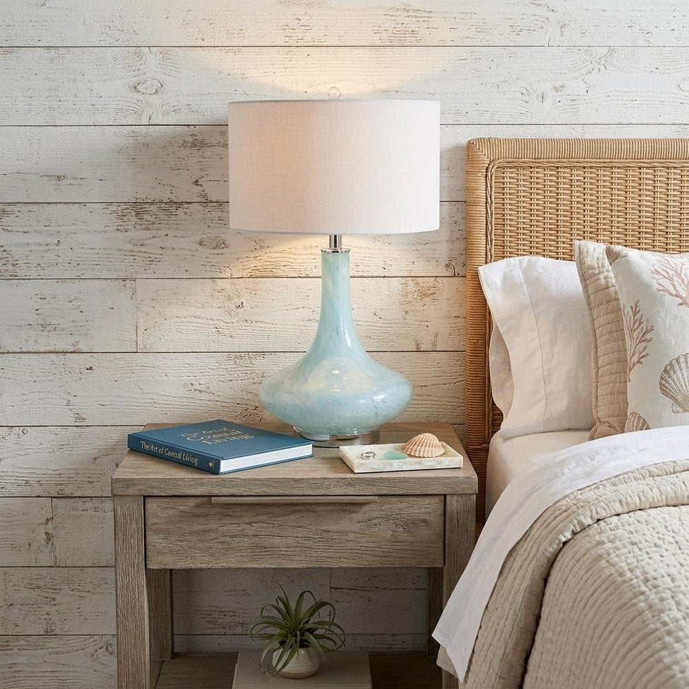 29" Light Blue And Clear Teardrop Glass Table Lamp With Drum Shade displayed on a dark wood tabletop in a furnished interior against an off-white wall.
