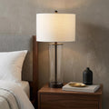 29" Antiqued Bronze Table Lamp With Drum Shade displayed on a dark wood tabletop in a furnished interior against a tan wall.