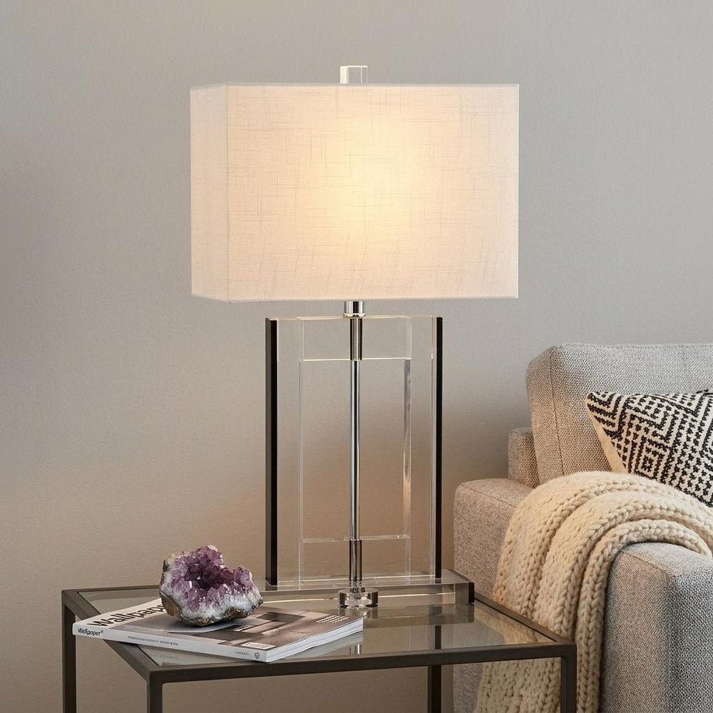 26" Clear Novelty Table Lamp With Drum Shade displayed on a dark wood tabletop in a furnished interior against a light gray wall.