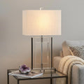 26" Clear Novelty Table Lamp With Drum Shade displayed on a dark wood tabletop in a furnished interior against a light gray wall.