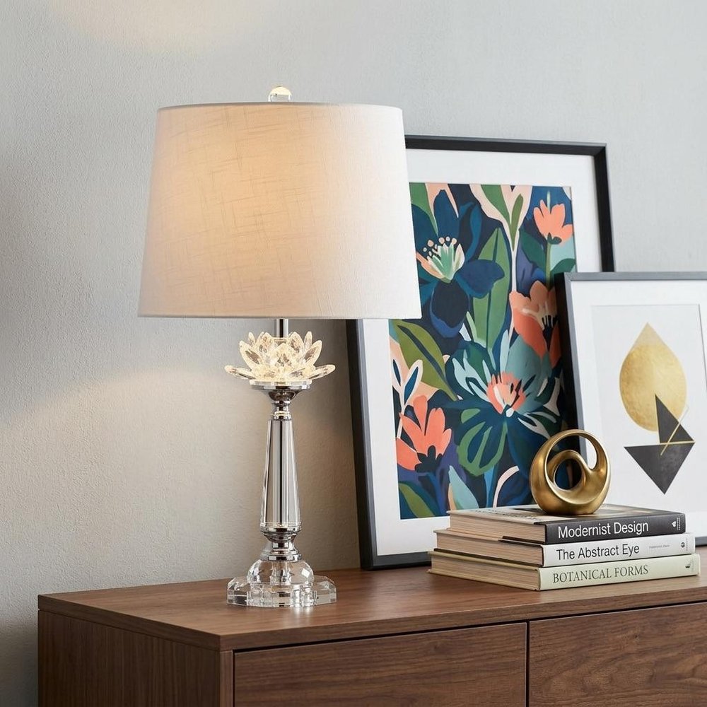 25" Clear Novelty Table Lamp With Drum Shade displayed on a dark wood tabletop in a furnished interior against a light gray wall.