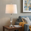 27" Silver Novelty Table Lamp With Empire Shade displayed on a dark wood tabletop in a furnished interior against a tan wall.