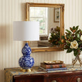 27" Blue Gold And White Novelty Table Lamp With Drum Shade displayed on a dark wood tabletop in a furnished interior against a cream wall.