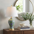 24" Green And Gold Urn Table Lamp With Empire Shade displayed on a dark wood tabletop in a furnished interior against an off-white wall.