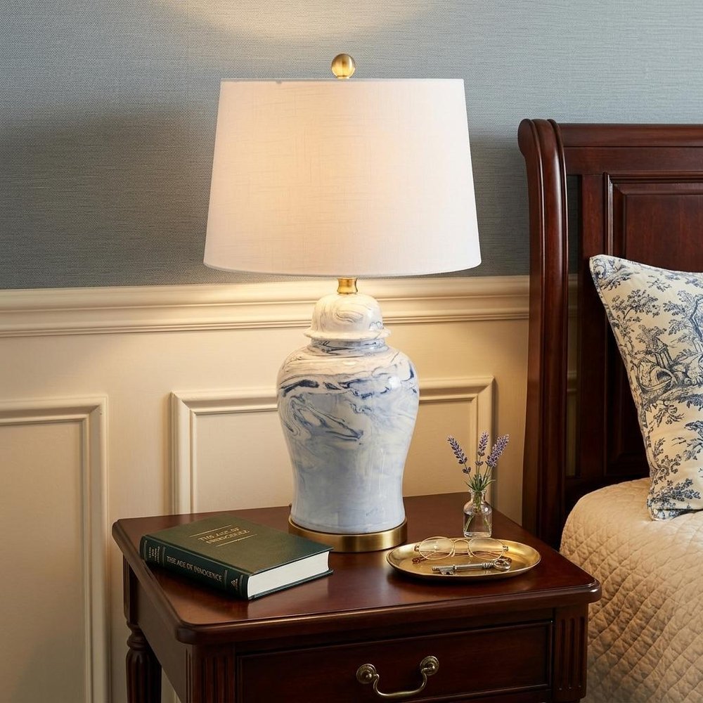 26" Gold Novelty Table Lamp With Drum Shade displayed on a tabletop in a furnished interior against a light gray wall.