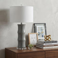 27" Gray And Clear Gourd Bedside Lamp With Drum Shade styled in a furnished interior with an off-white wall on a dark wood floor.