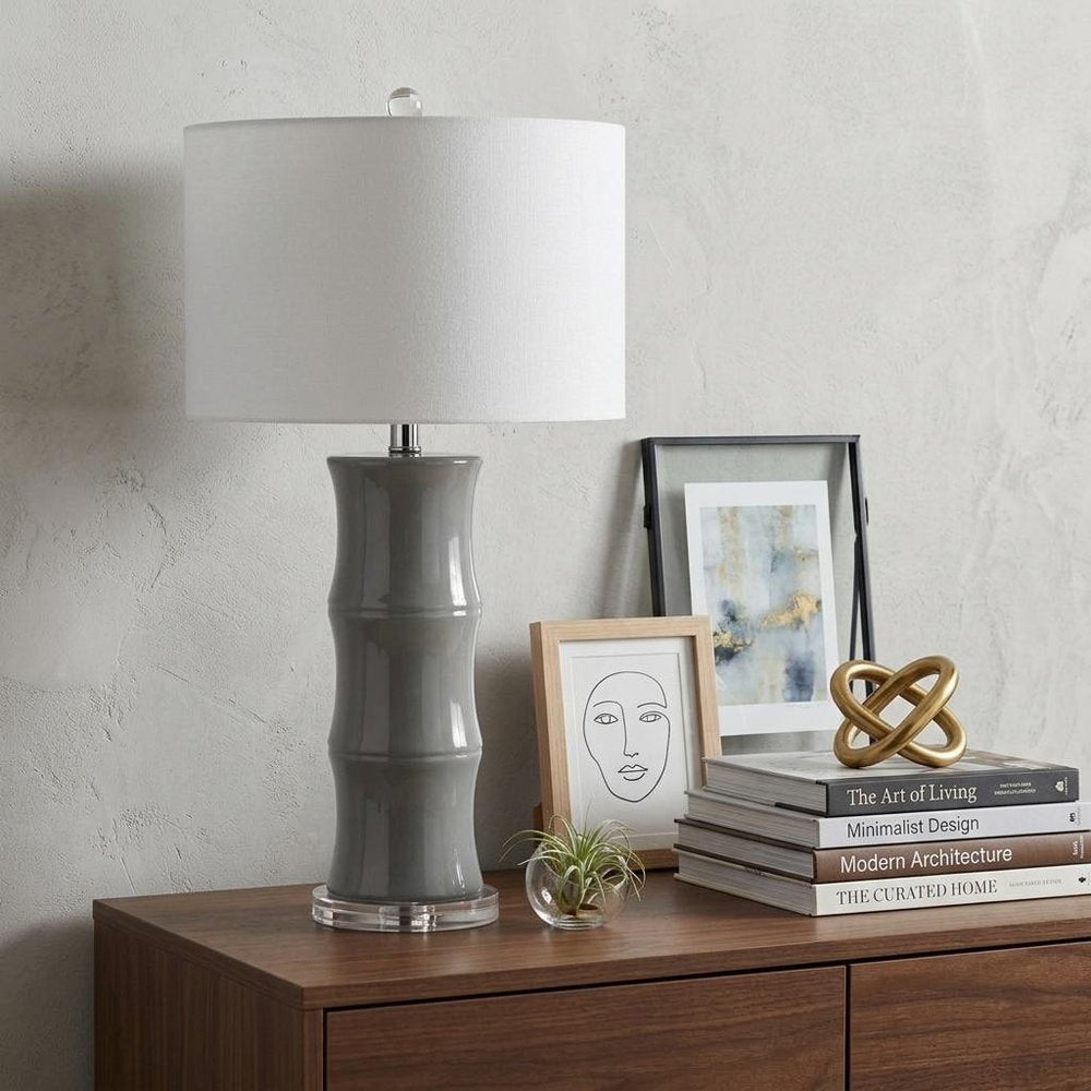 27" Gray And Clear Gourd Bedside Lamp With Drum Shade styled in a furnished interior with an off-white wall on a dark wood floor.