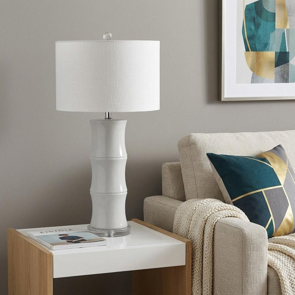 27" Clear Table Lamp With Drum Shade displayed on a dark wood tabletop in a furnished interior against a light gray wall.