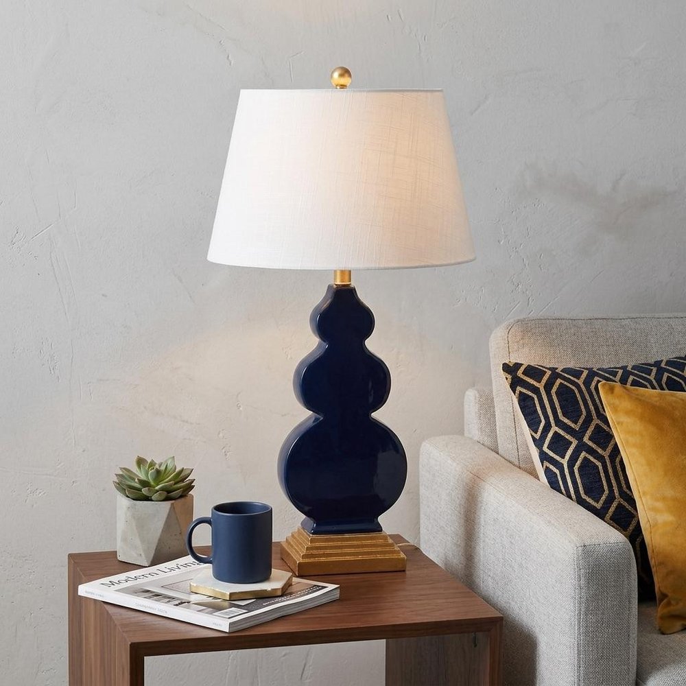 29" Gold Novelty Table Lamp With Drum Shade displayed on a dark wood tabletop in a furnished interior against a light gray wall.