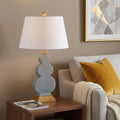 29" Gold Novelty Table Lamp With Drum Shade displayed on a dark wood tabletop in a furnished interior against a tan wall.