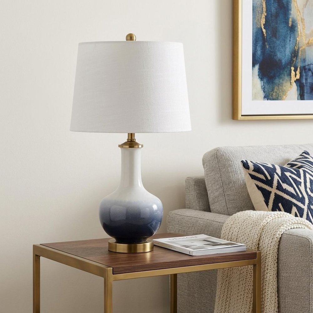 25" Gold Novelty Table Lamp With Drum Shade displayed on a wood tabletop in a furnished interior against an off-white wall.