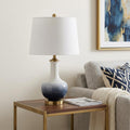 25" Gold Novelty Table Lamp With Drum Shade displayed on a wood tabletop in a furnished interior against an off-white wall.