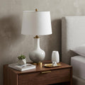 25" Gold Novelty Table Lamp With Drum Shade displayed on a dark wood tabletop in a furnished interior against a light gray wall.