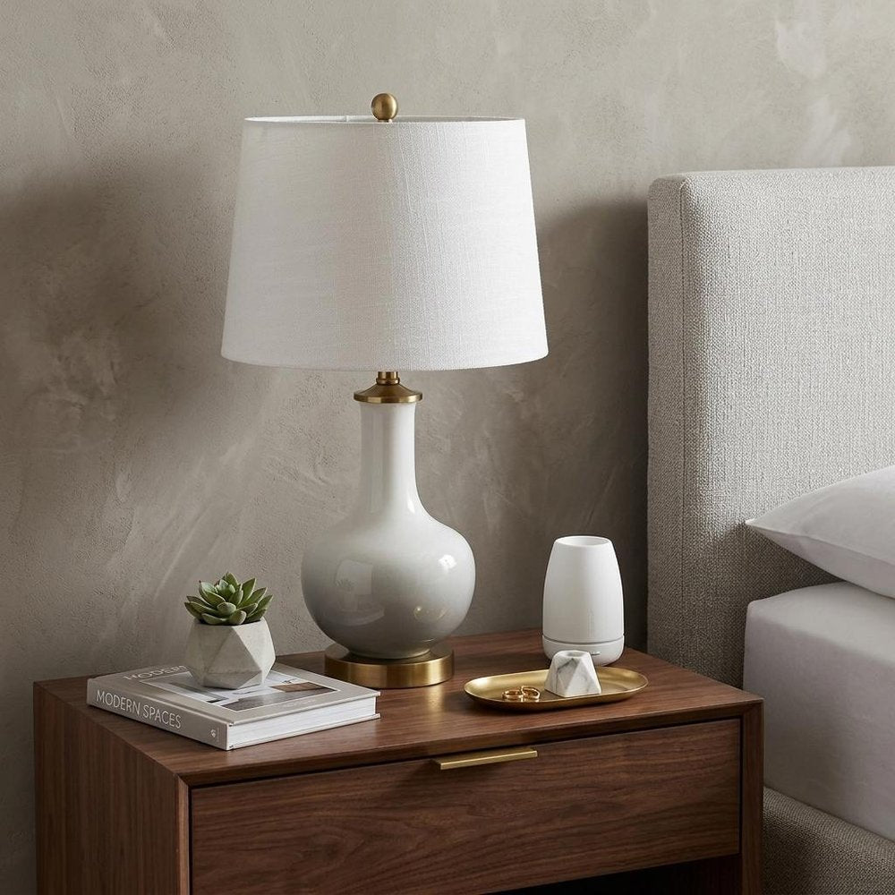 25" Gold Novelty Table Lamp With Drum Shade displayed on a dark wood tabletop in a furnished interior against a light gray wall.