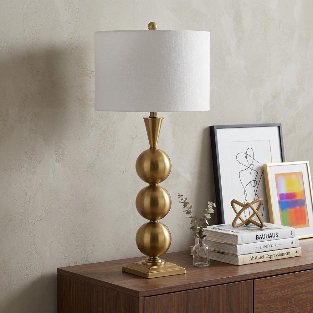 33" Gold Novelty Table Lamp With Drum Shade displayed on a dark wood tabletop in a furnished interior against a light gray wall.