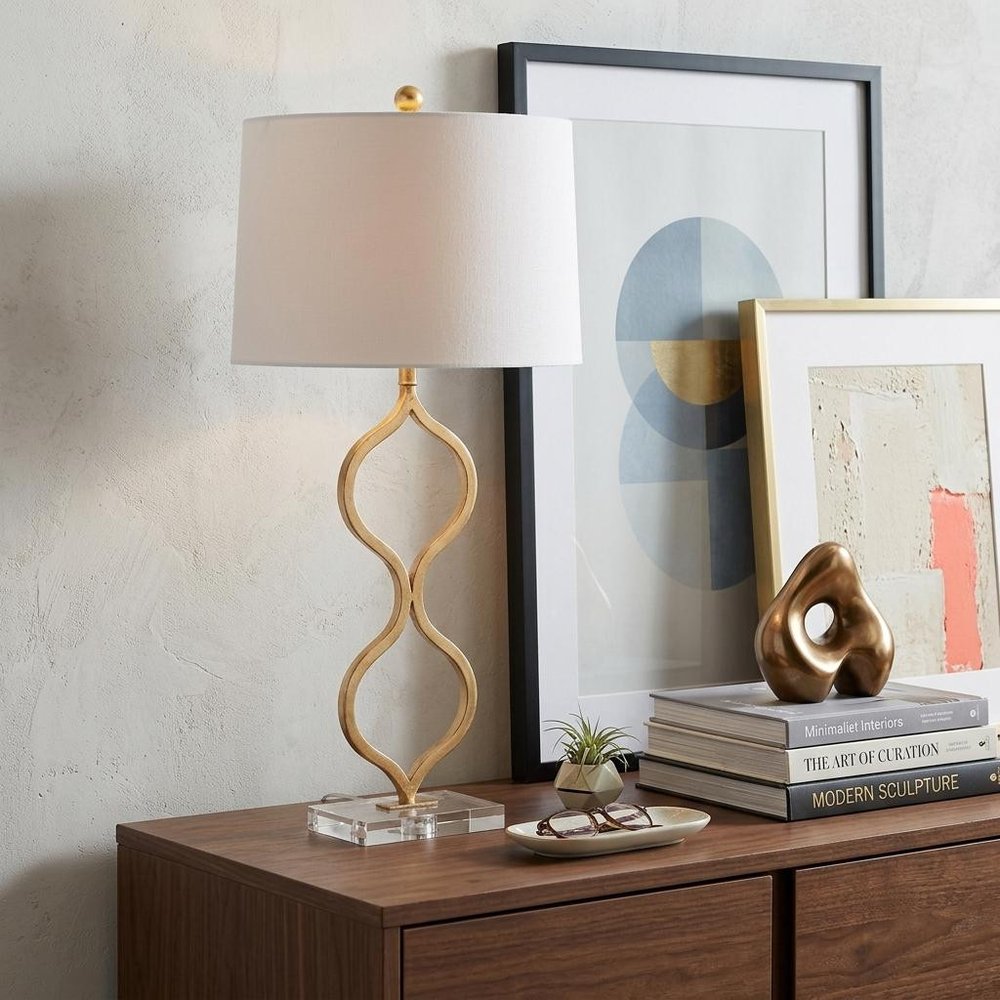 32" Clear Novelty Table Lamp With Drum Shade displayed on a dark wood tabletop in a furnished interior against an off-white wall.