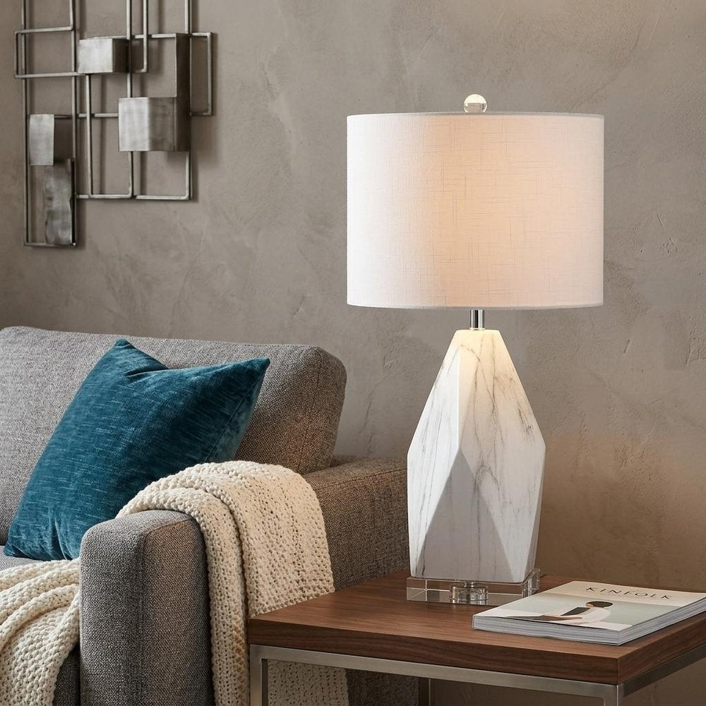 26" Clear Novelty Table Lamp With Drum Shade displayed on a dark wood tabletop in a furnished interior against a tan wall.
