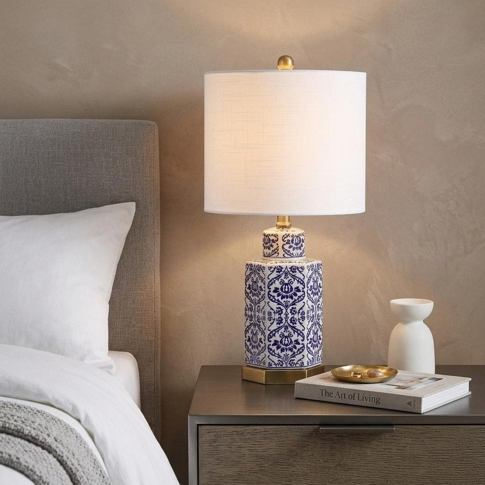24" Navy Blue Gold And White Table Lamp With Drum Shade displayed on a dark wood tabletop in a furnished interior against a tan wall.
