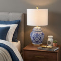 25" Gold Novelty Table Lamp With Drum Shade displayed on a dark wood tabletop in a furnished interior against a gray wall.