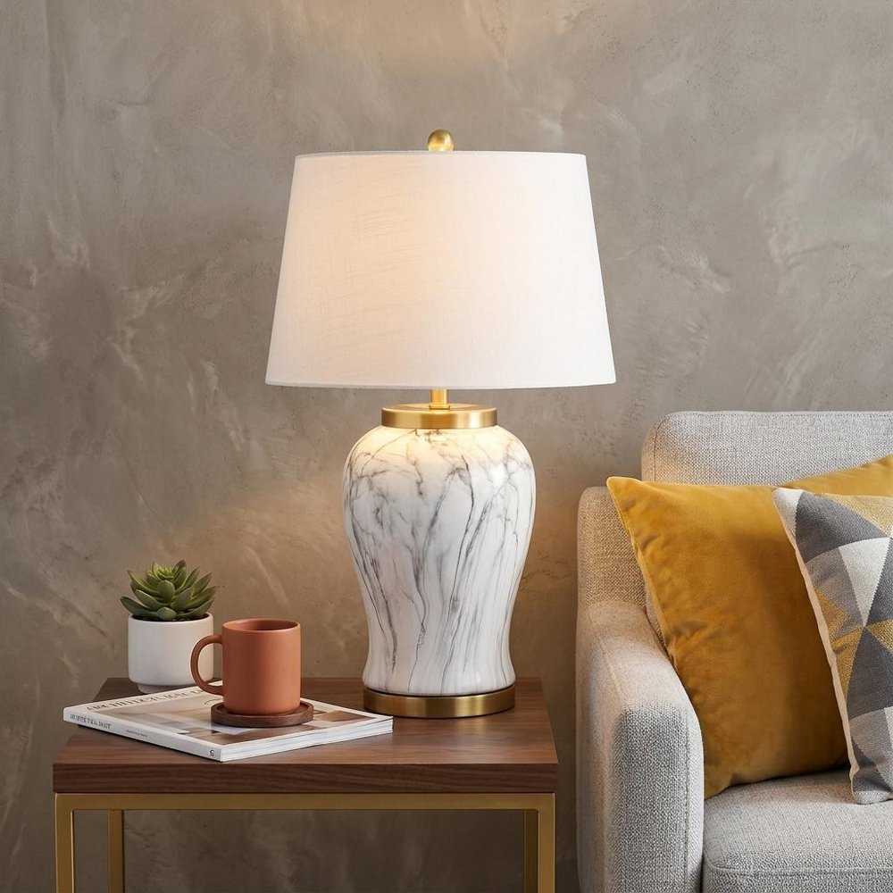 28" Gray White And Gold Urn Table Lamp With Empire Shade displayed on a dark wood tabletop in a furnished interior against a tan wall.