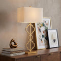 32" Gold Geometric Table Lamp With Shade displayed on a dark wood tabletop in a furnished interior against a tan wall.