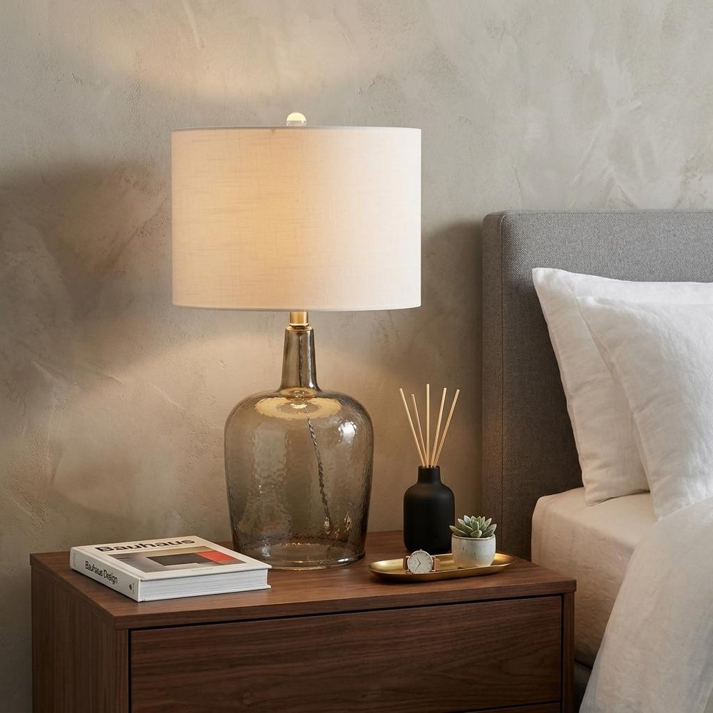 26" Clear Urn Table Lamp With Drum Shade displayed on a dark wood tabletop in a furnished interior against a tan wall.