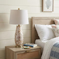 28" Gold Novelty Table Lamp With Drum Shade displayed on a dark wood tabletop in a furnished interior against an off-white wall.