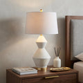 29" White And Gold Novelty Bedside Lamp With Drum Shade styled in a furnished interior with a light gray wall on a dark wood floor.