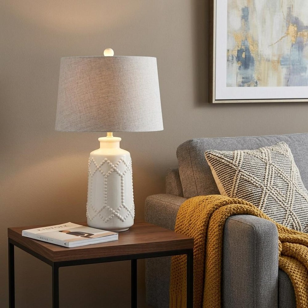 26" Cream Table Lamp With Drum Shade displayed on a dark wood tabletop in a furnished interior against a tan wall.