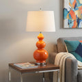 28" Orange And Silver Novelty Table Lamp With Drum Shade displayed on a dark wood tabletop in a furnished interior against a light gray wall.