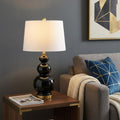 28" Gold Novelty Table Lamp With Drum Shade displayed on a dark wood tabletop in a furnished interior against a gray wall.