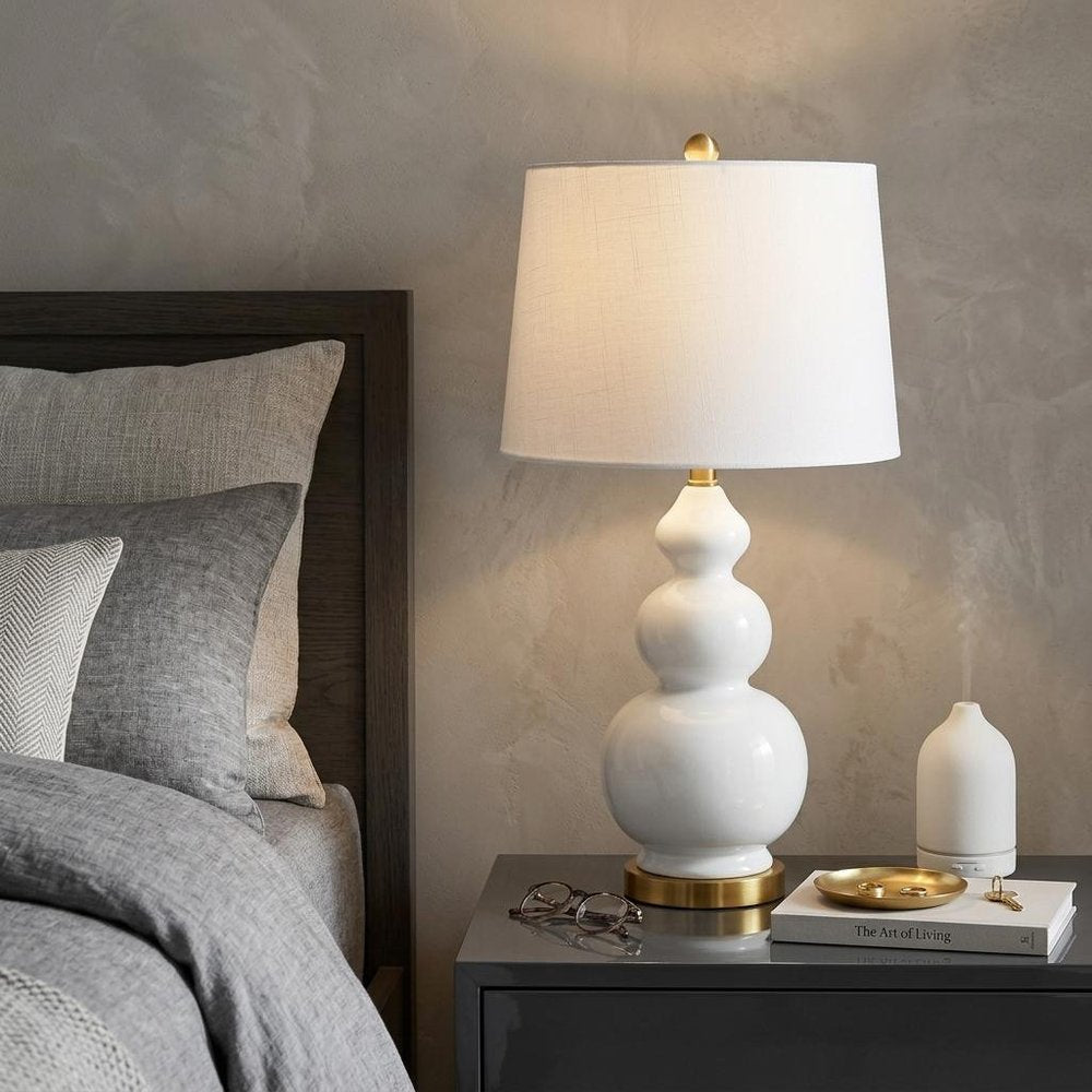 28" Gold Novelty Table Lamp With Drum Shade displayed on a tabletop in a furnished interior against a tan wall.
